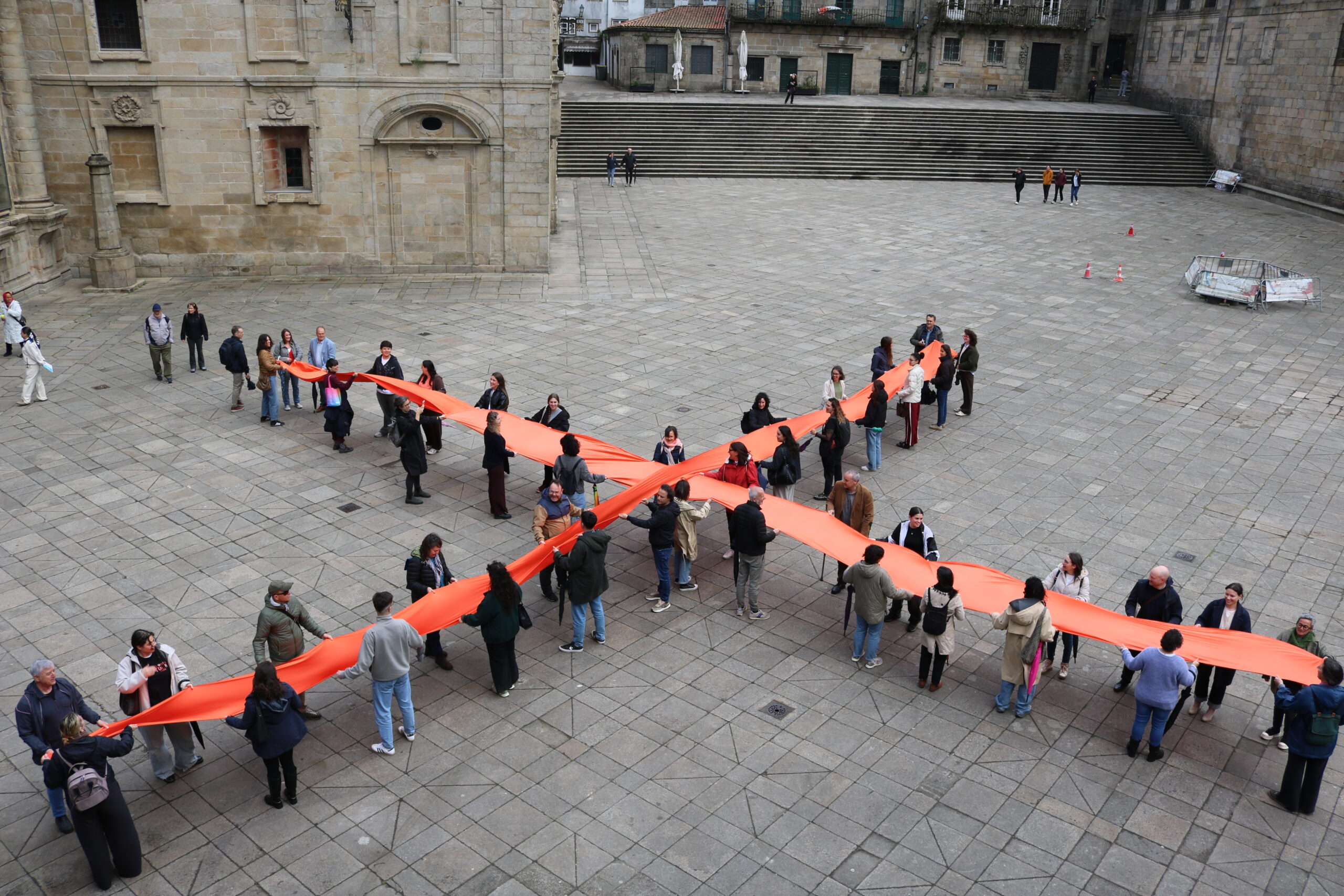 Participantes de una campaña solidaria sosteniendo telas naranjas cruzadas en una plaza histórica de piedra.